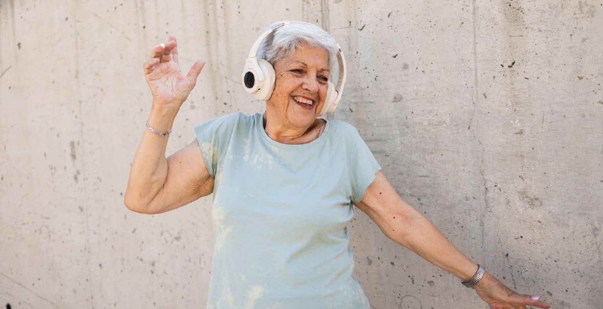 Elderly woman with short gray hair wearing headphones, smiling and dancing against a concrete wall.