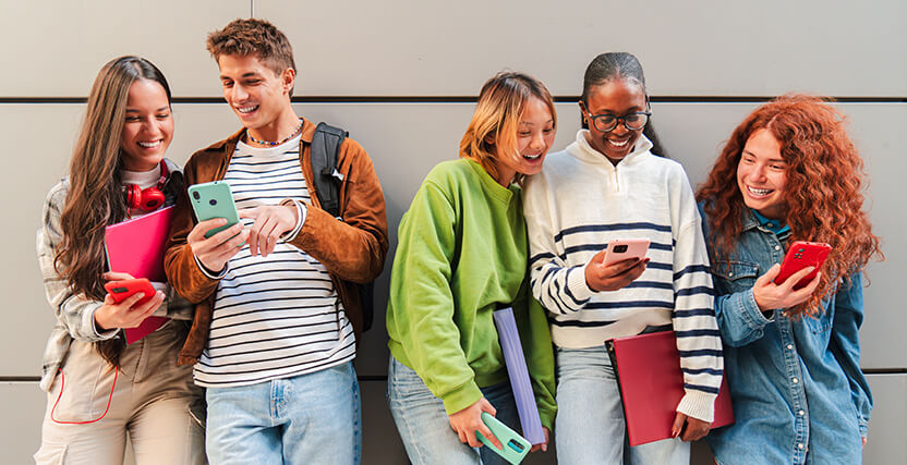 Five young adults smiling and looking at their smartphones, standing against a wall. They hold books and wear casual clothing.