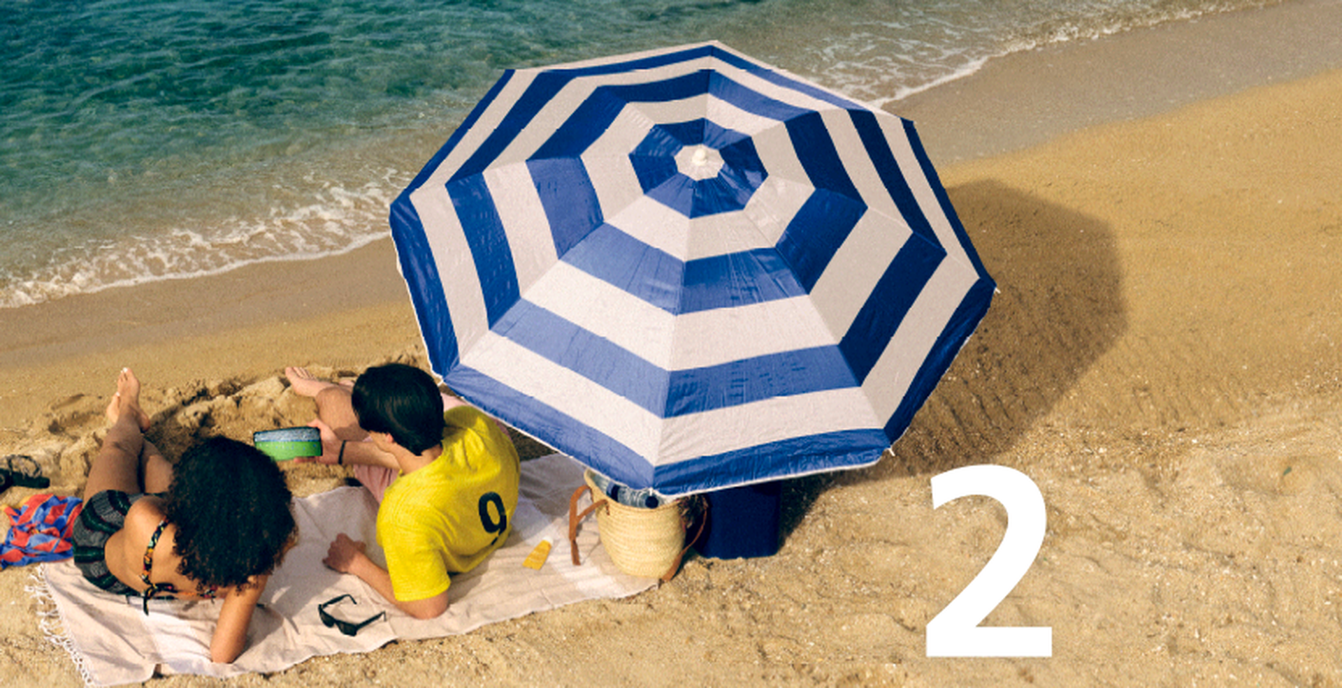 Two people relaxing under a blue and white striped umbrella on a sandy beach with a small number "2" on the sand.