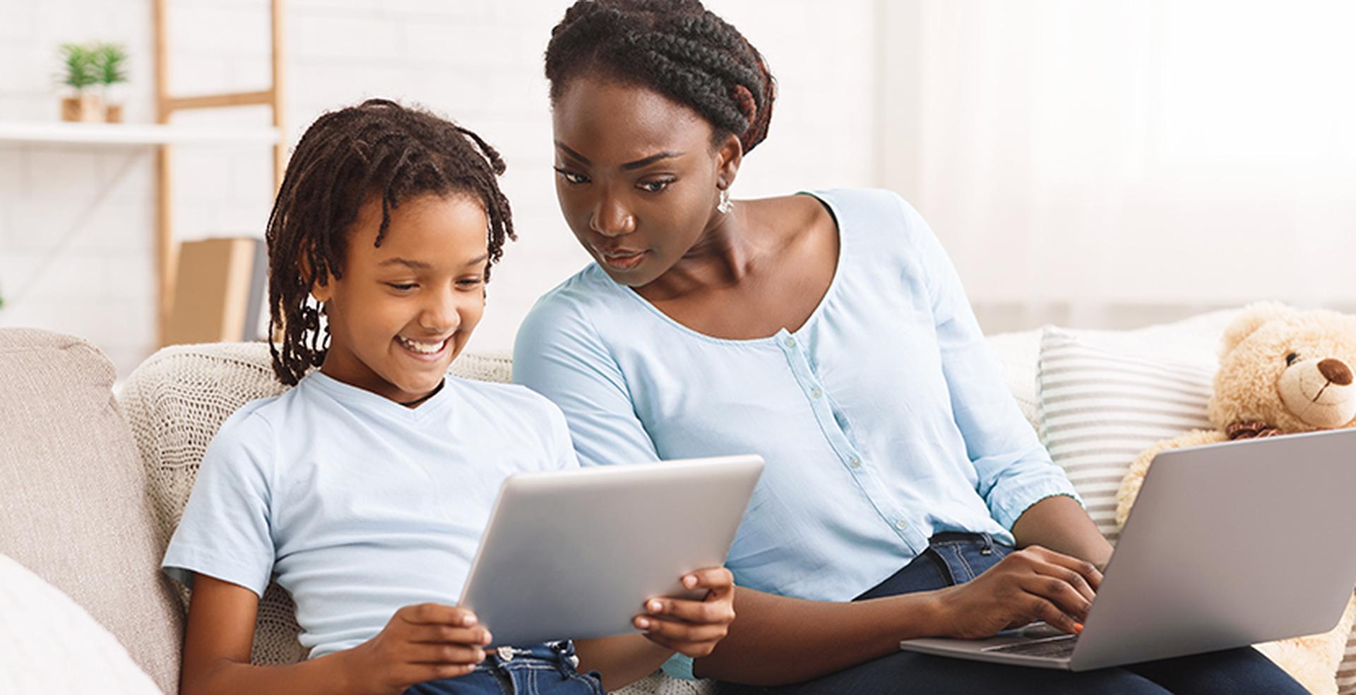 Woman and child sitting on a couch, both wearing light blue shirts. The child is using a tablet, and the woman is using a laptop.