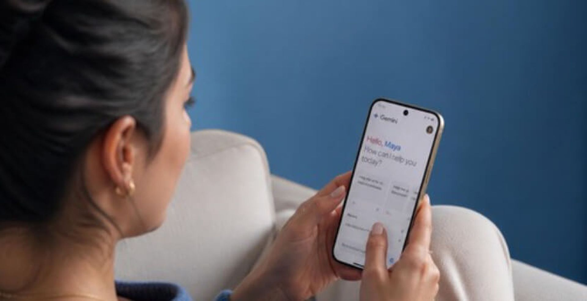 A woman sitting on a couch holds a smartphone displaying an app interface with the text, "Hello, Maya. How can I help you today?"