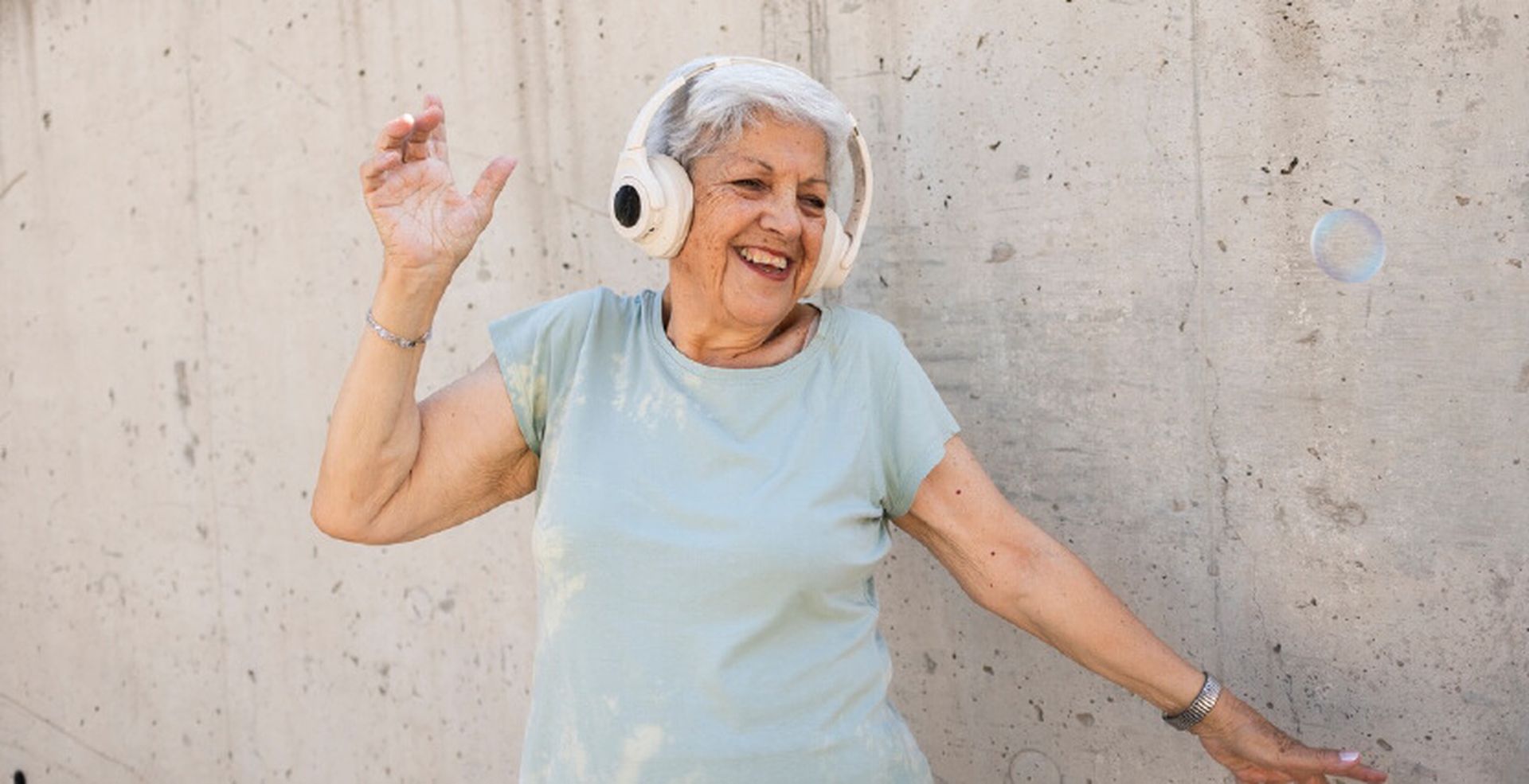 Elderly woman joyfully dancing with headphones on, smiling against a concrete wall background.