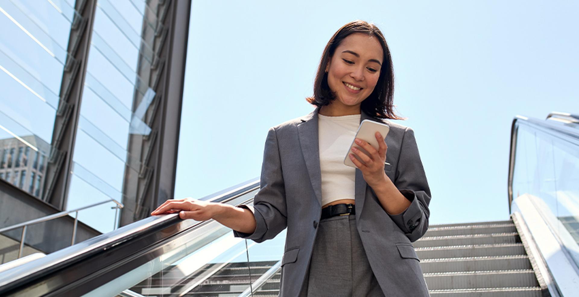 Woman in a gray suit smiles while looking at her phone, standing on an outdoor escalator against a clear blue sky.