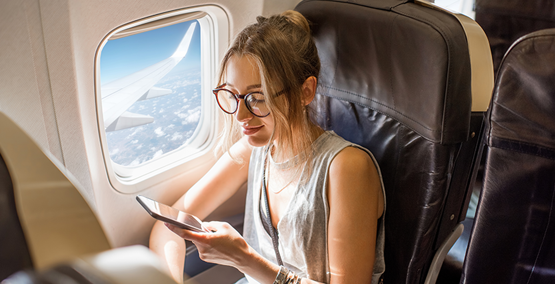 Woman with glasses using a smartphone, sitting by an airplane window. Bright daylight outside with a view of the wing and clouds.