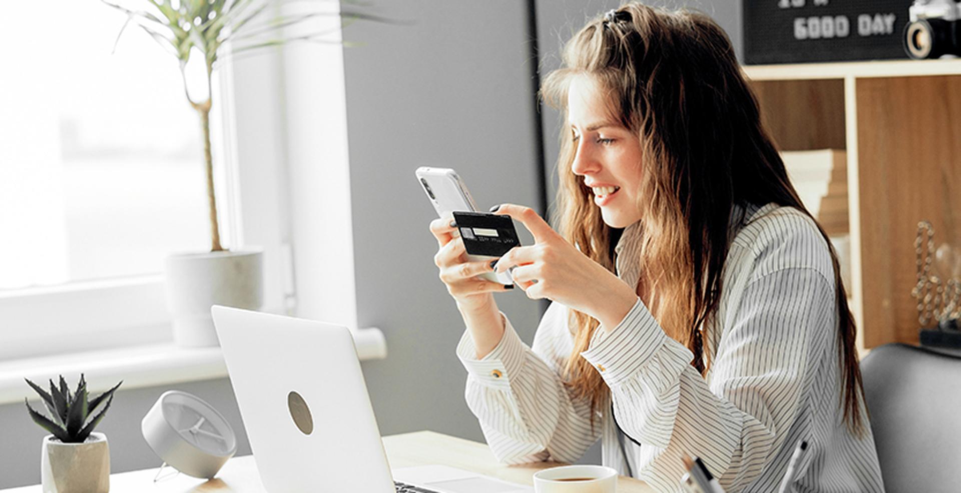 Woman at desk with a smartphone and credit card, smiling. Laptop, plant, and cup beside her; window light brightens the scene.