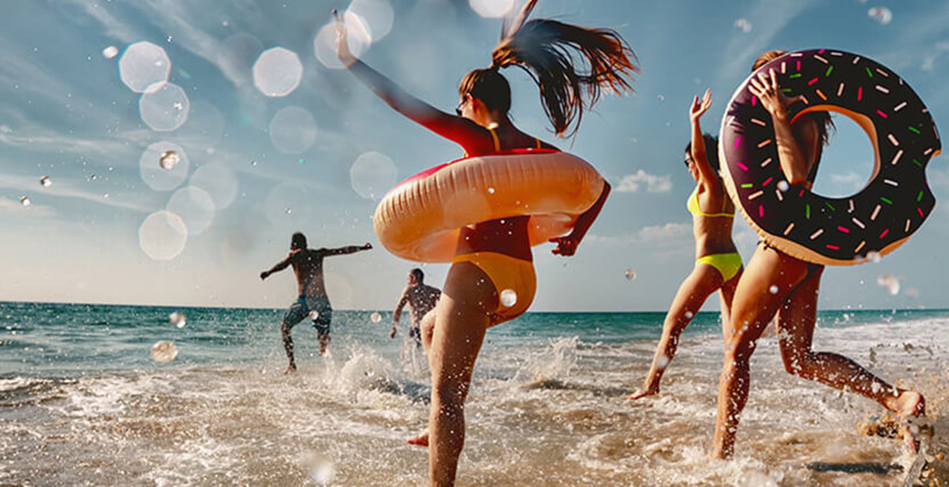 People joyfully playing in the ocean surf, wearing swimsuits and inflatable rings, with sunlight and water droplets creating a vibrant, energetic scene.