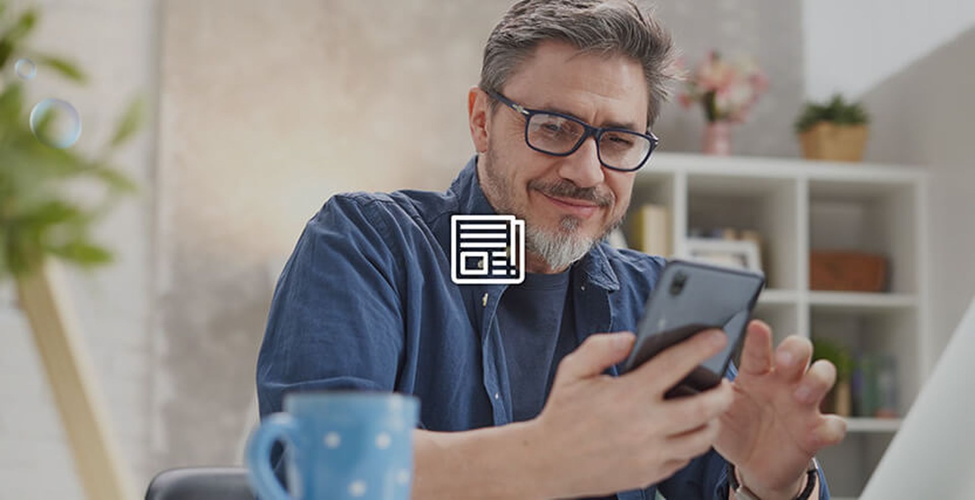 Man with glasses smiling while looking at a smartphone. Blue mug on the table, books and plants on the bookshelf in the background.