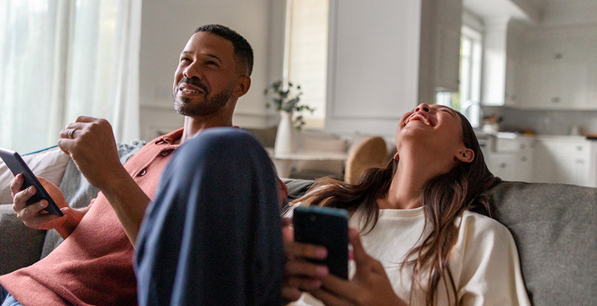 A man and woman sitting on a couch, smiling and using their smartphones, in a bright, modern living room.