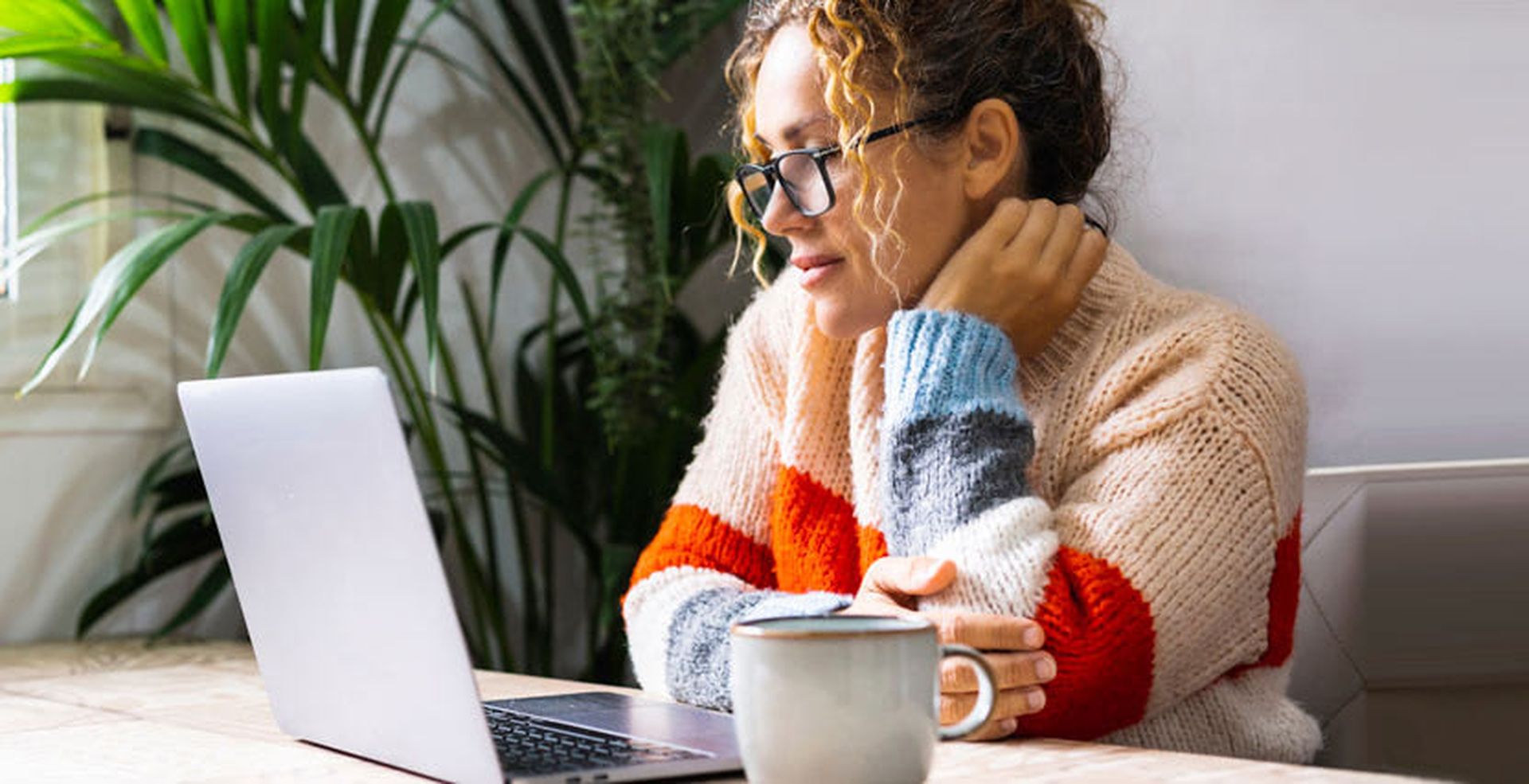 A person with glasses and curly hair sits at a table, looking at a laptop, with a mug nearby and plants in the background.