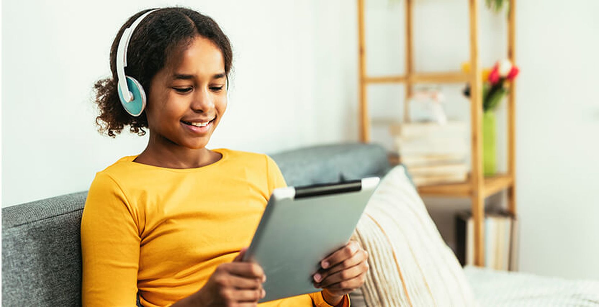 A young teen with headphones smiles while using a tablet on a sofa, with books and flowers in the background.