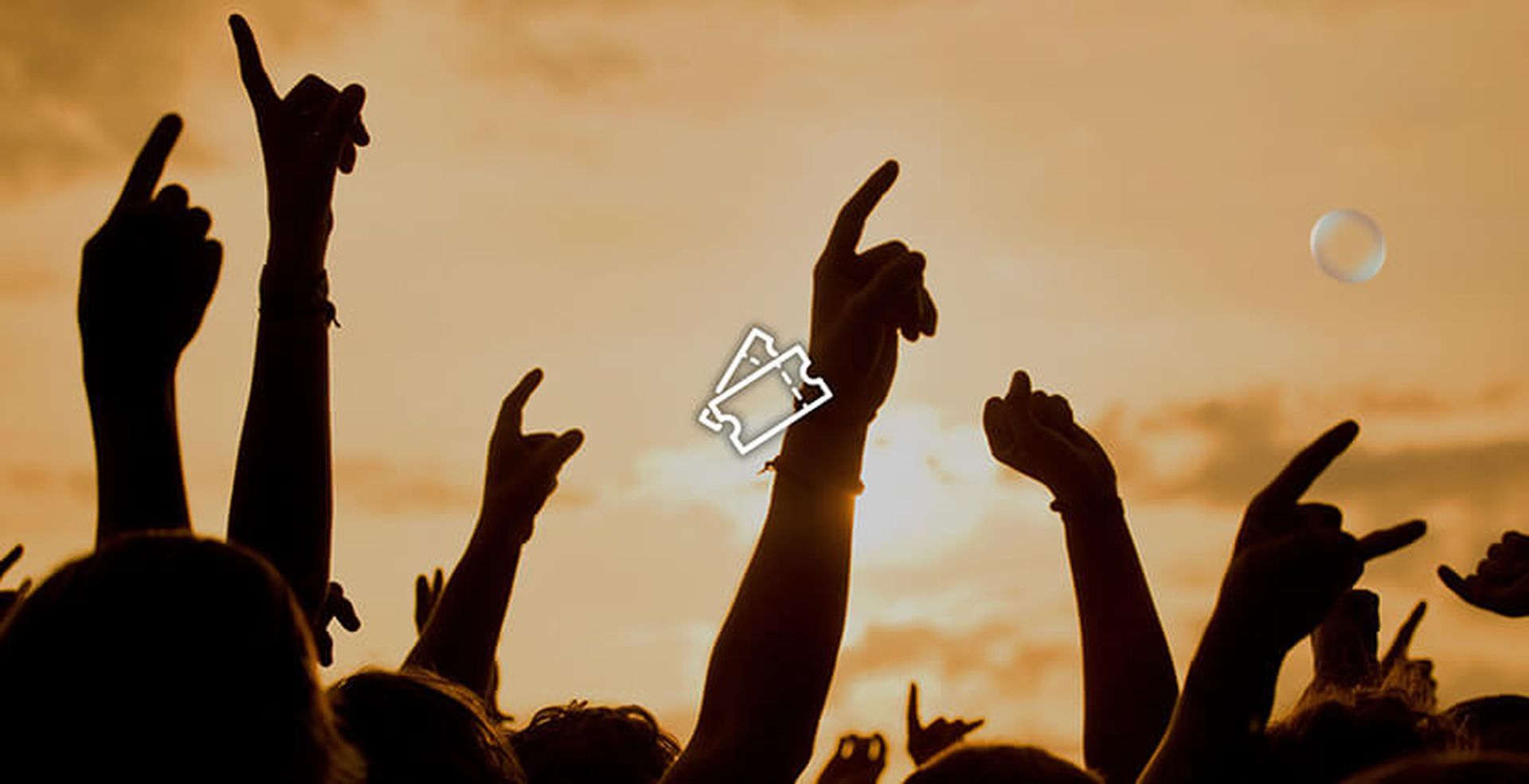 Silhouettes of raised hands at a concert against an orange sunset sky.