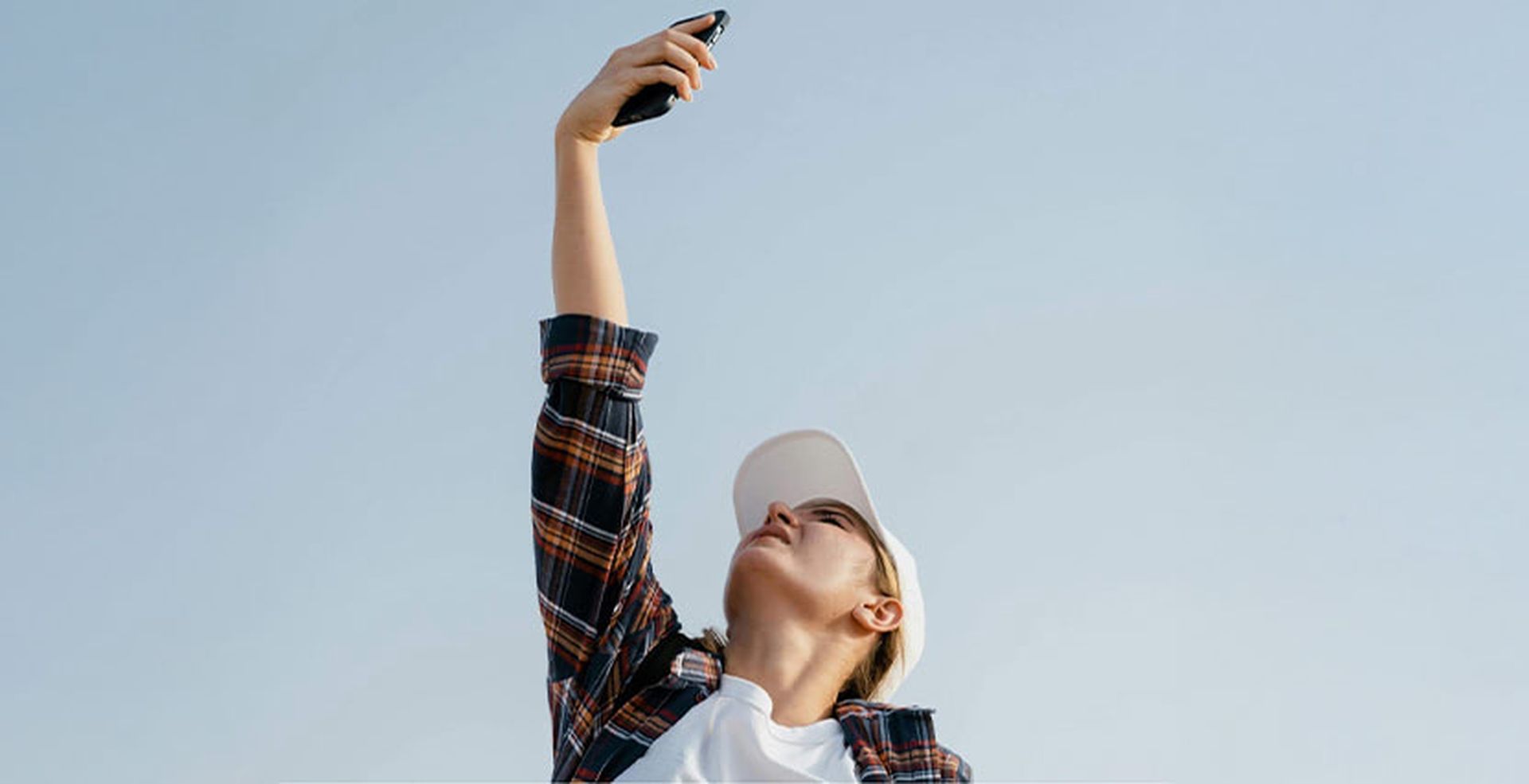 Person in a plaid shirt and cap takes a selfie with a smartphone held high against a clear blue sky.