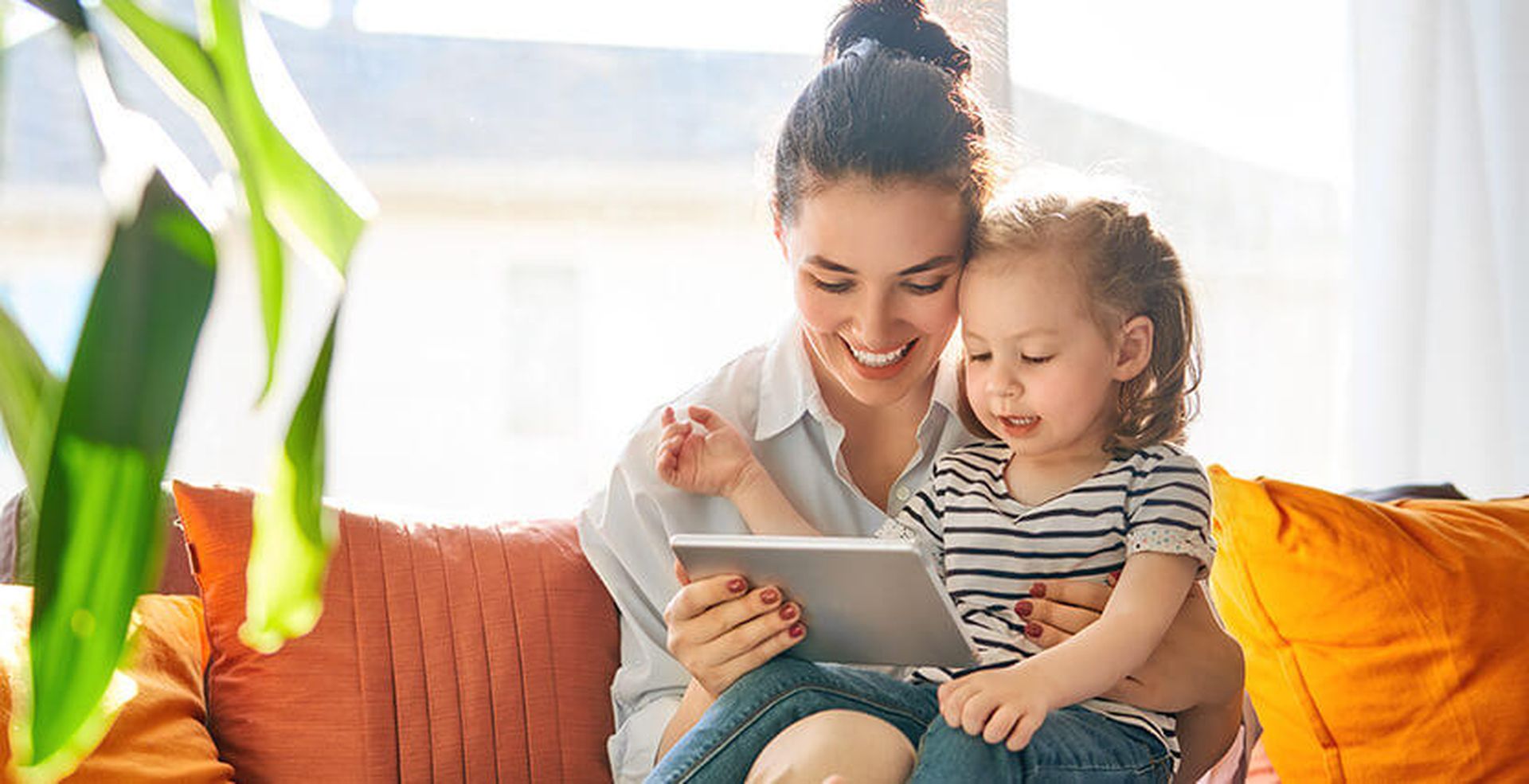 A woman and child sit on a sofa, smiling and looking at a tablet. Sunlight streams through a window, illuminating the room with orange cushions.