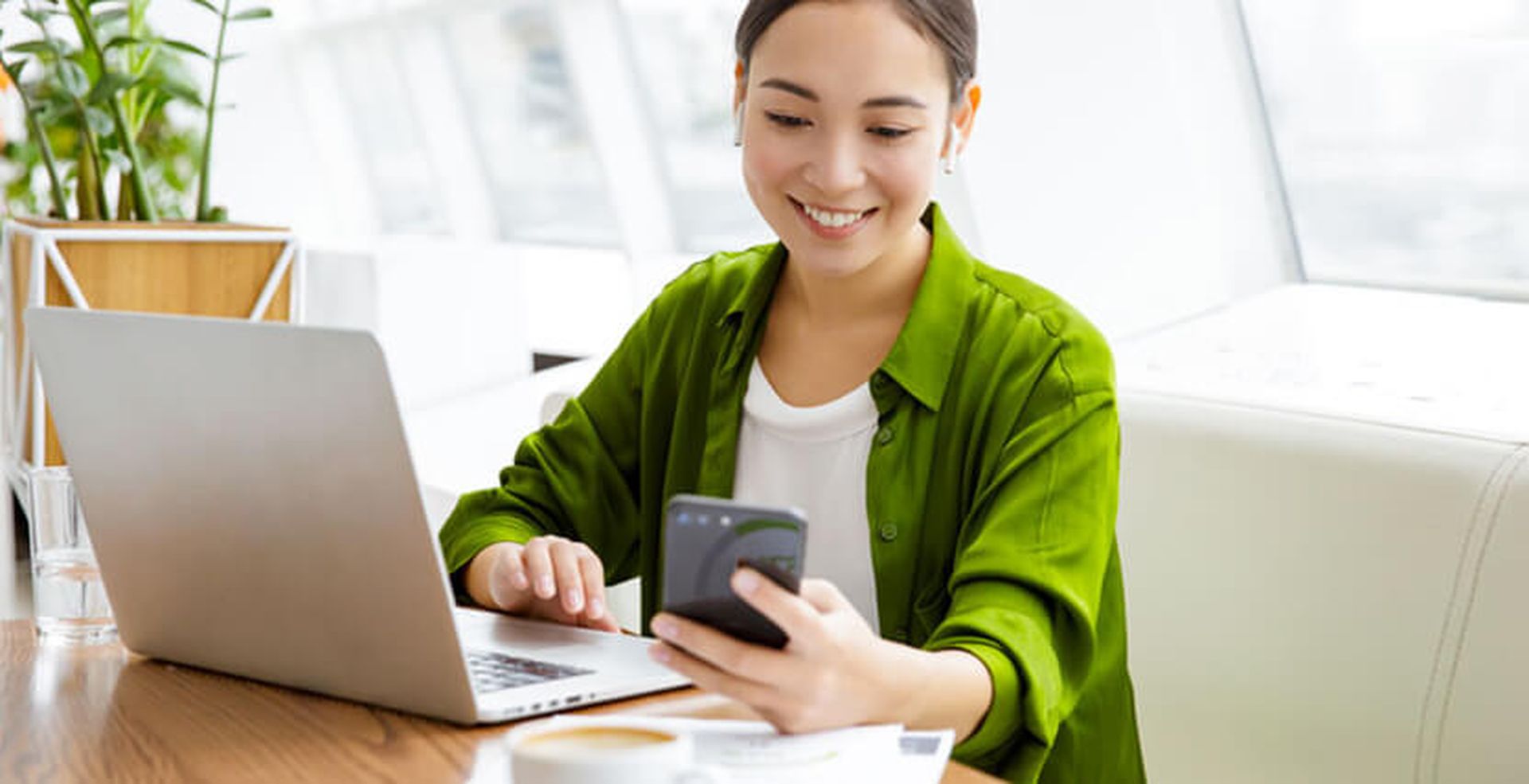 Smiling woman in green shirt using smartphone and laptop at a desk with coffee, bright office setting with plants in the background.