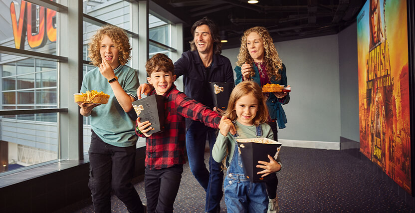A family of five excitedly walks through a theater hallway, holding popcorn and snacks, with movie posters on the walls.