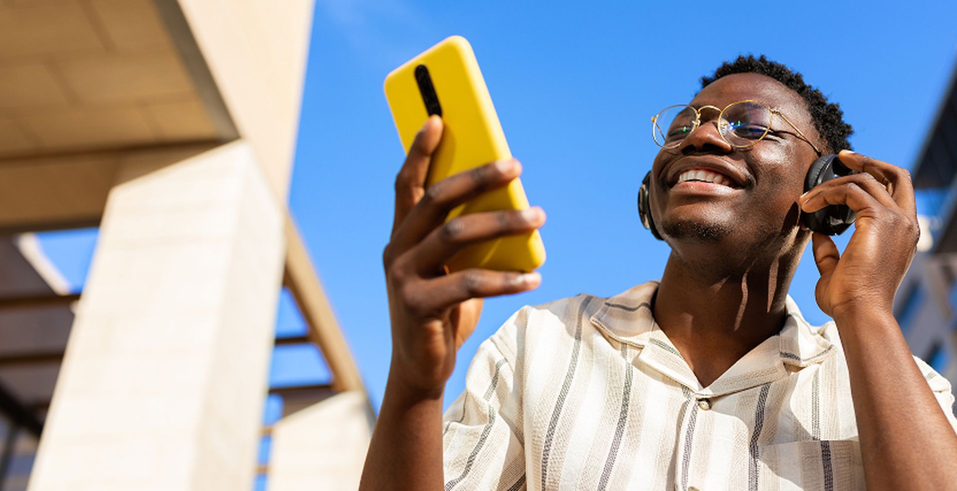 A smiling person with headphones uses a yellow smartphone outdoors, wearing a striped shirt and glasses, under a clear blue sky.