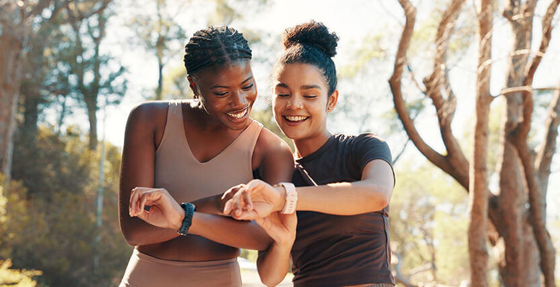 Two women smiling, checking fitness watches outdoors in a wooded area, wearing athletic clothing.