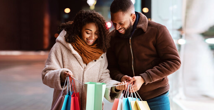 A smiling couple in winter coats happily looks at colorful shopping bags while standing on a city street at night.