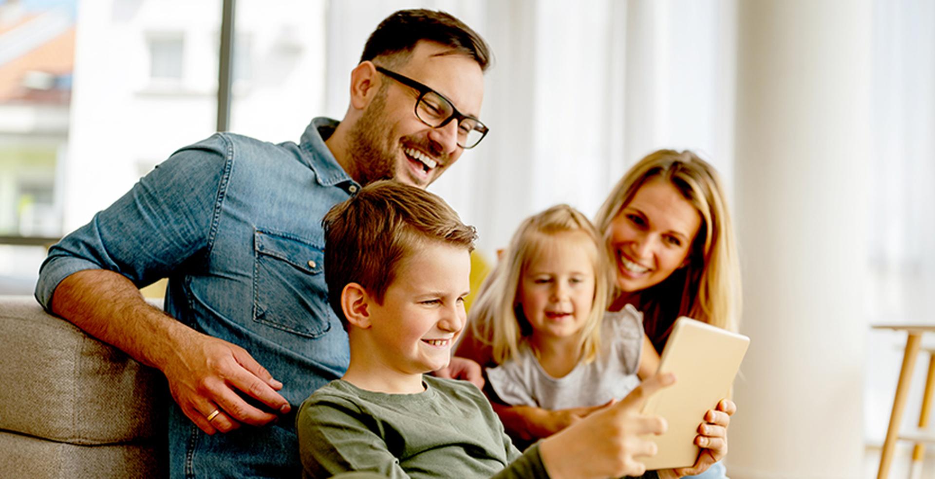 A smiling family of four, including a young boy and girl, sitting on a couch, looking at a tablet.