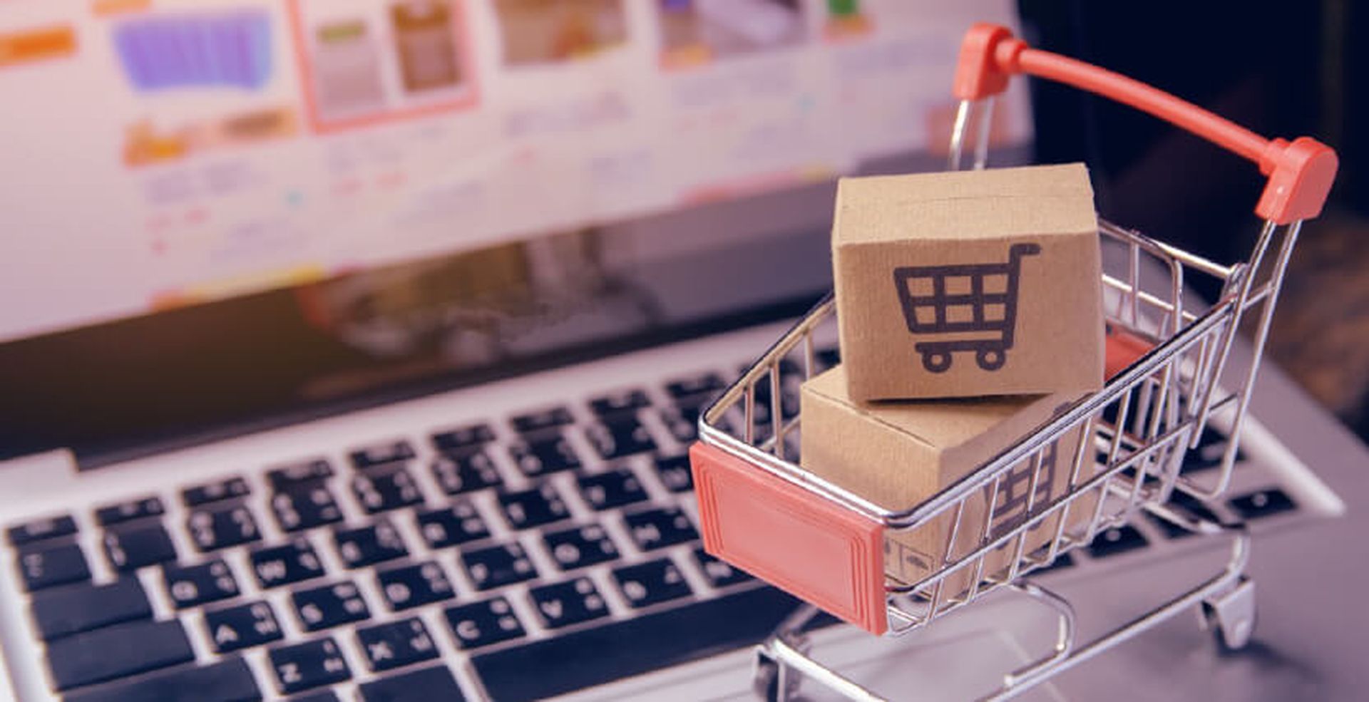 A miniature shopping cart with cardboard boxes sits on a laptop keyboard, symbolizing online shopping.