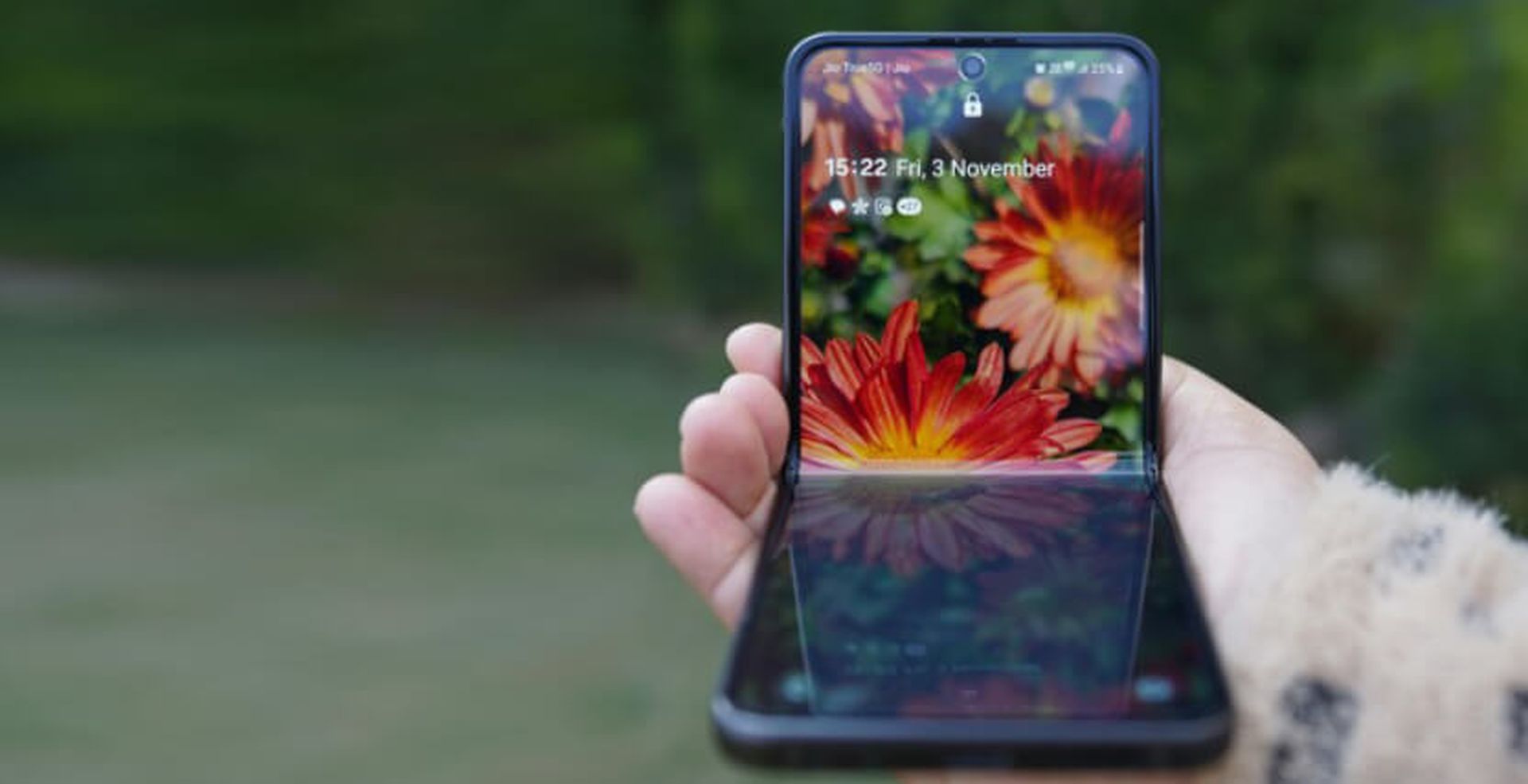 A person holds a smartphone with a floral wallpaper and the time displayed as 15:22 on Friday, 3 November. The phone's screen reflects sunlight.