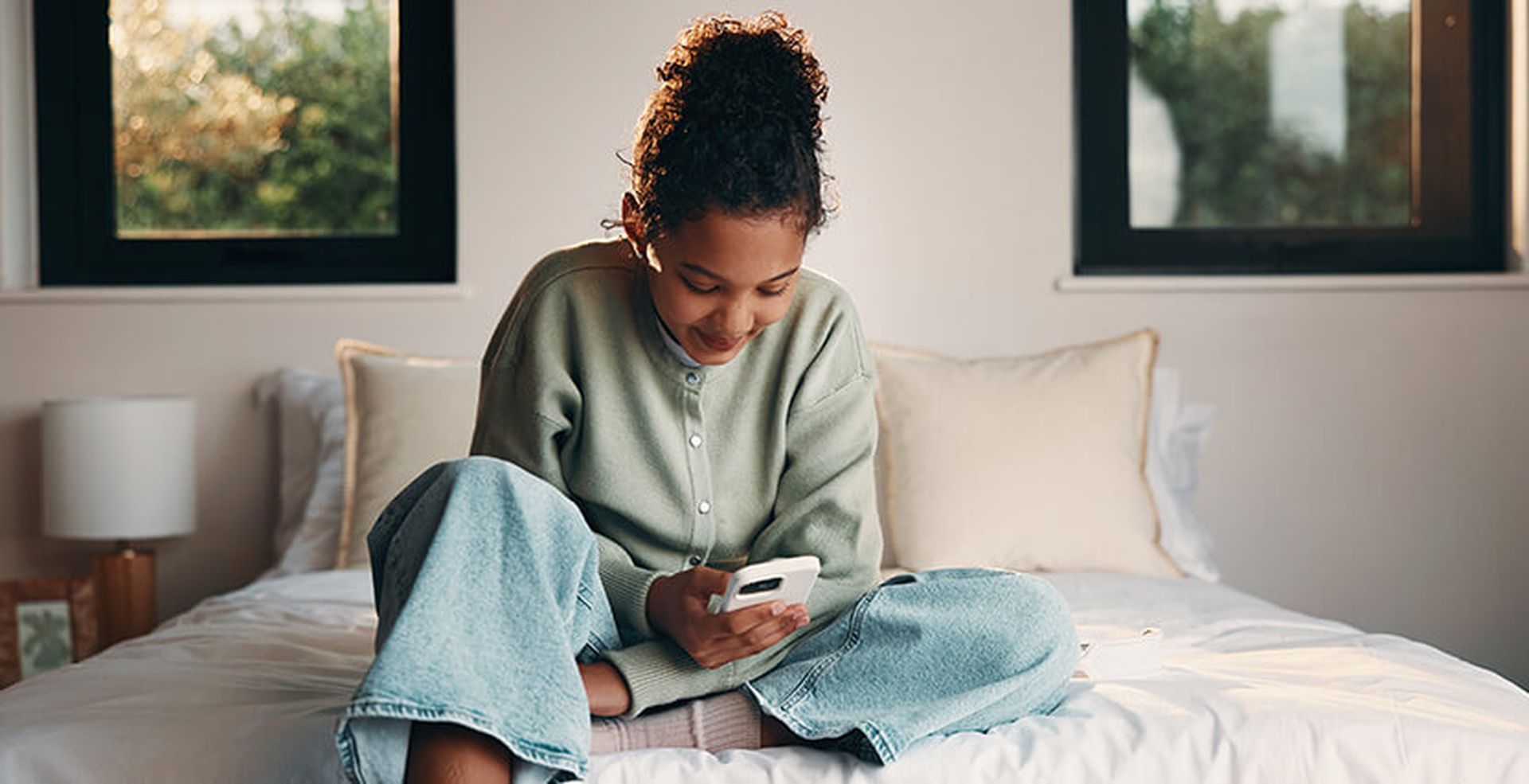 Person smiling while looking at a phone, sitting cross-legged on a bed with white linens, in a room with two windows.