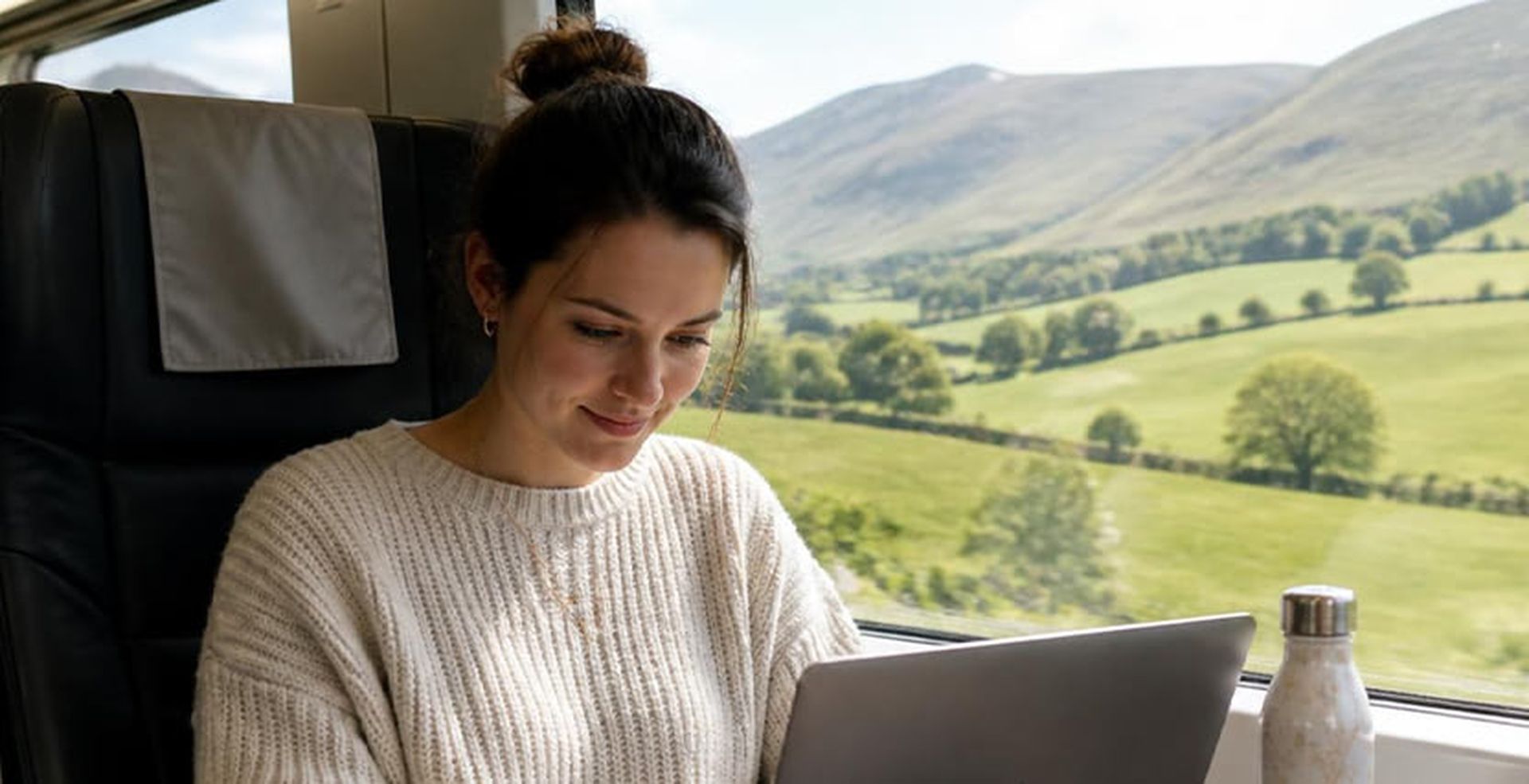 A woman in a white sweater works on her laptop while sitting on a train, with scenic green hills visible through the window.