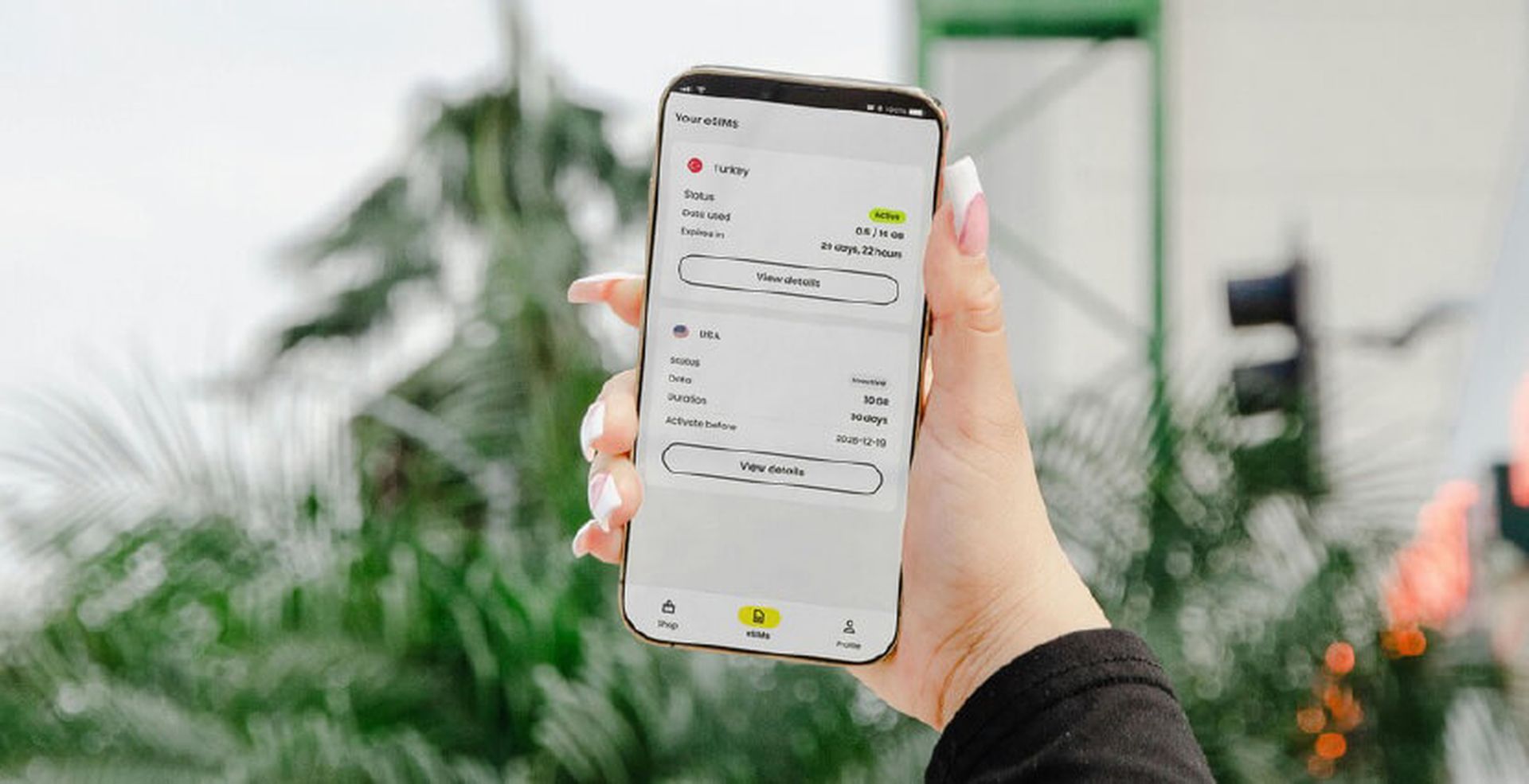 A hand holding a smartphone showing a data usage app with details on eSIMs. The background features blurred greenery and an urban setting.