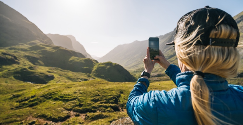 A person in a blue jacket and cap takes a photo of a sunlit, mountainous landscape with a smartphone.
