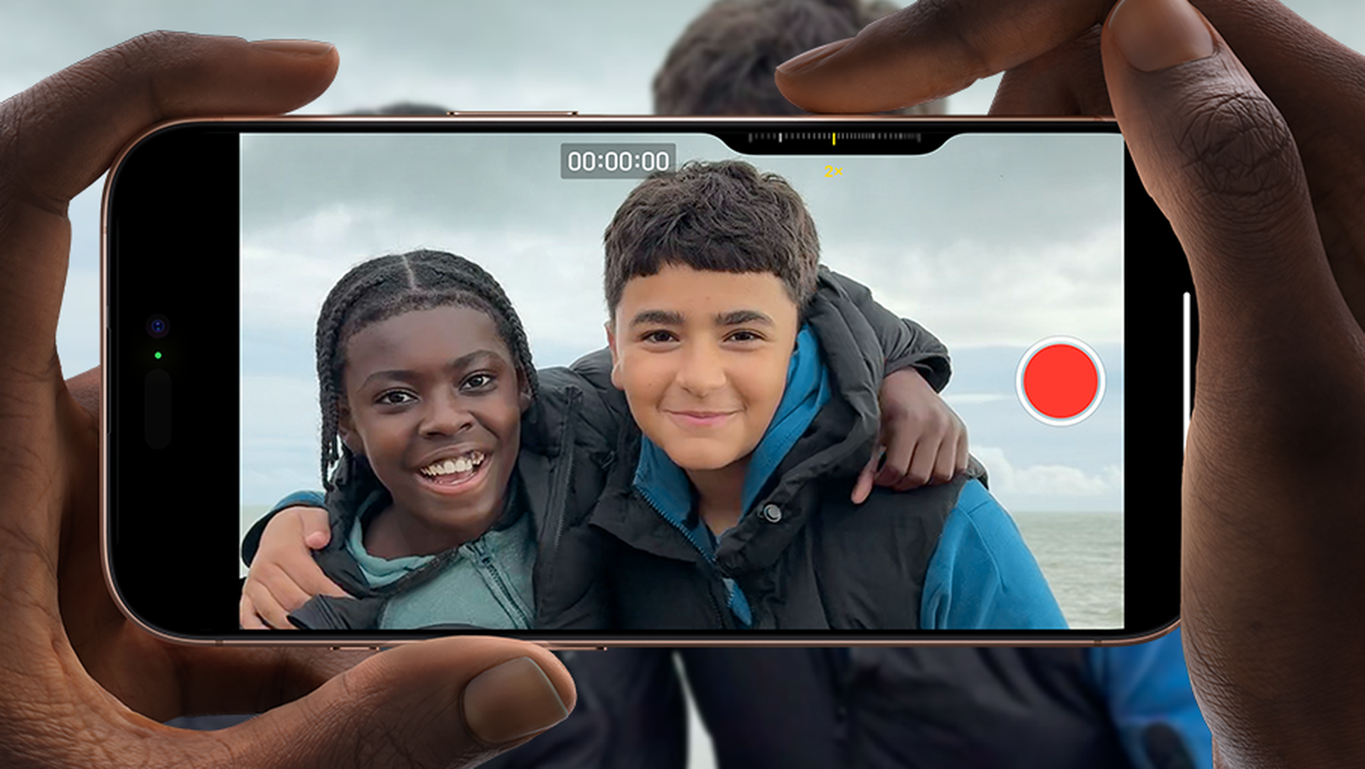 Two friends smiling at the camera, with one arm around each other, are being recorded on a smartphone by a person holding it.