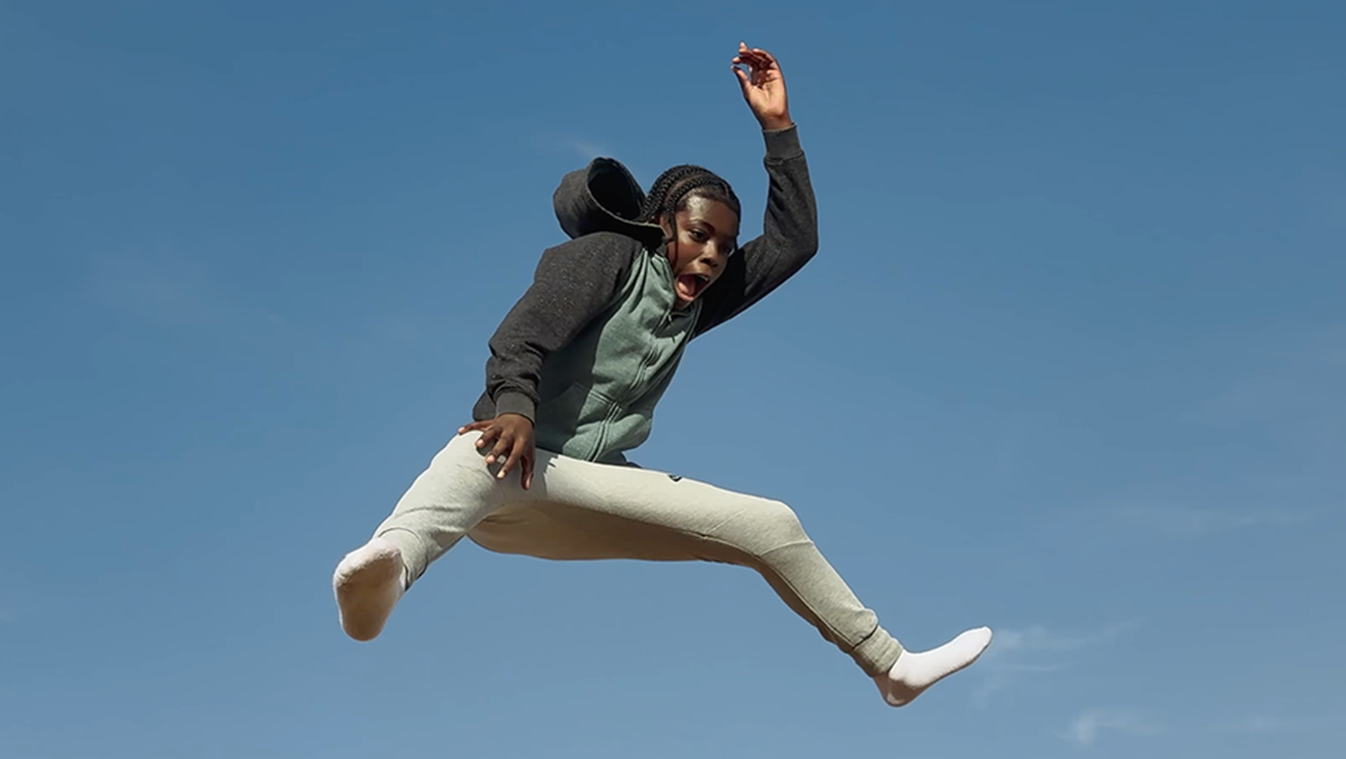 A person in mid-air against a clear blue sky, wearing a hoodie and light pants, with arms and legs outstretched in a dynamic pose.